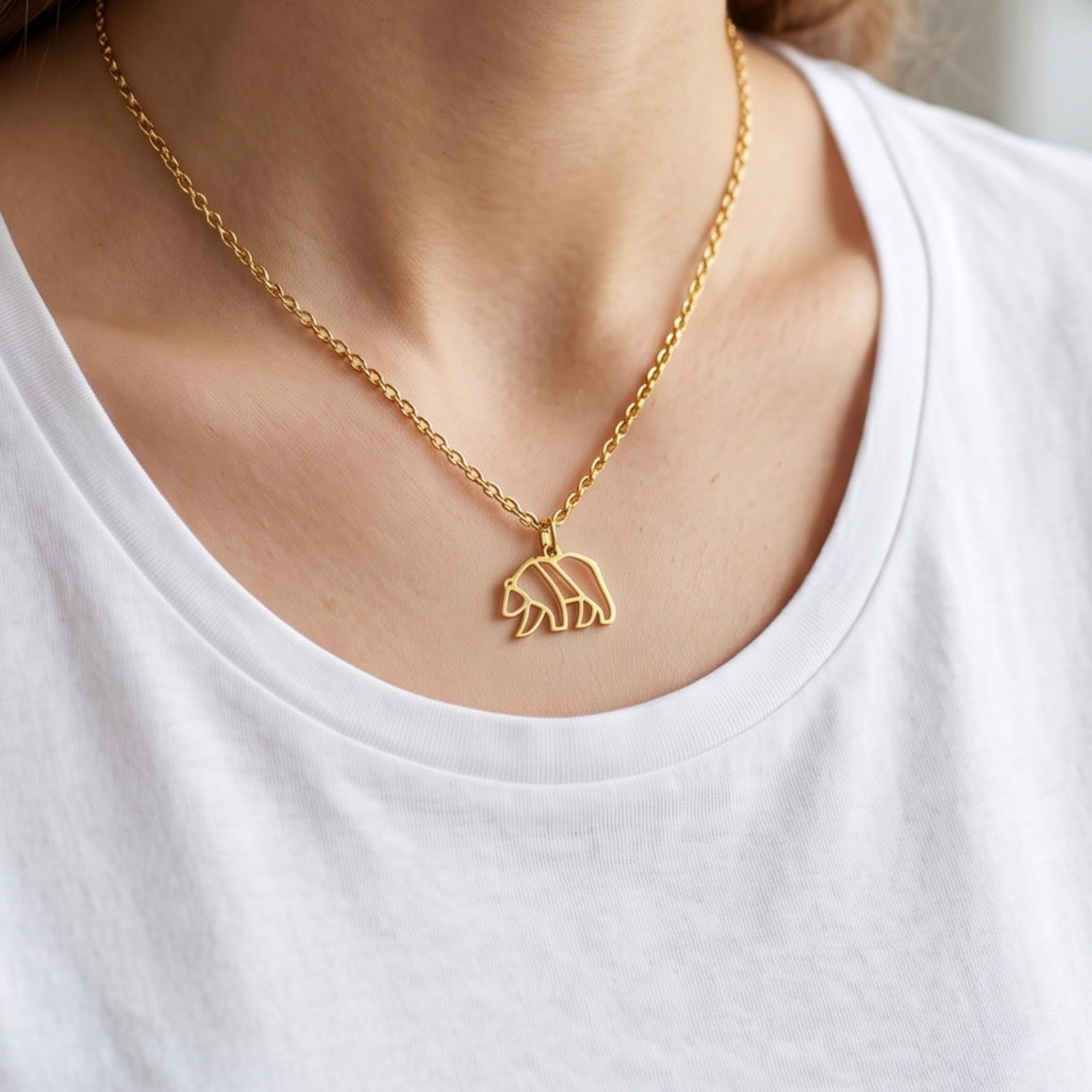 Gros plan sur un collier minimaliste en or porté par une femme, avec un pendentif géométrique et filaire en forme d'ours marchant, sur un tee-shirt blanc.