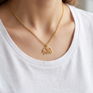 Gros plan sur un collier minimaliste en or porté par une femme, avec un pendentif géométrique et filaire en forme d'ours marchant, sur un tee-shirt blanc.