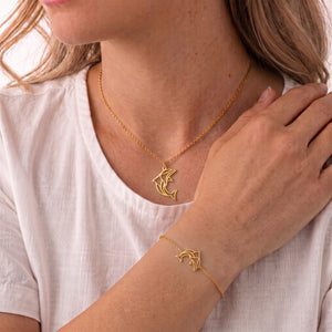 Parure de bijoux dauphin animal totem LIYAMI Nature : collier et bracelet dorés en acier inoxydable avec pendentifs dauphin géométrique, portés par une femme.